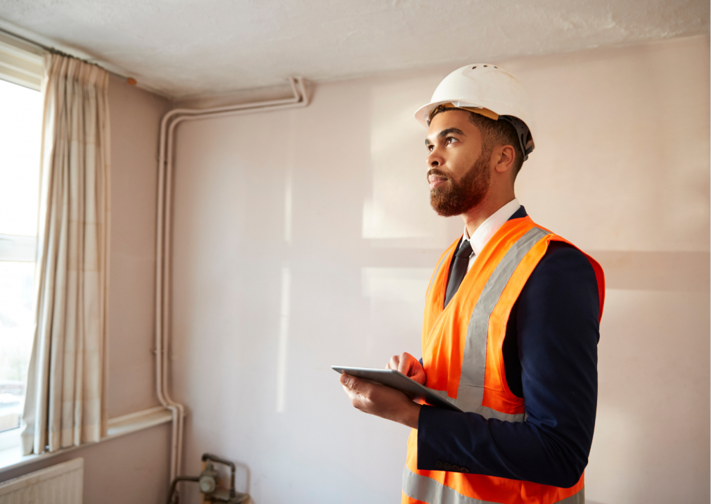 A man wearing a high vis vest and a hard hat, holding a clipboard, inspects the internal of a property to check for damage.