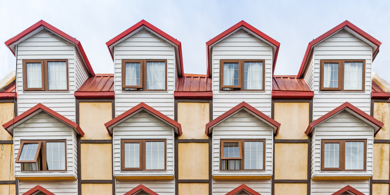 Image shows the top of a building with a red pitched roof. It is very symmetrical with white wooden siding and dark wood windows.