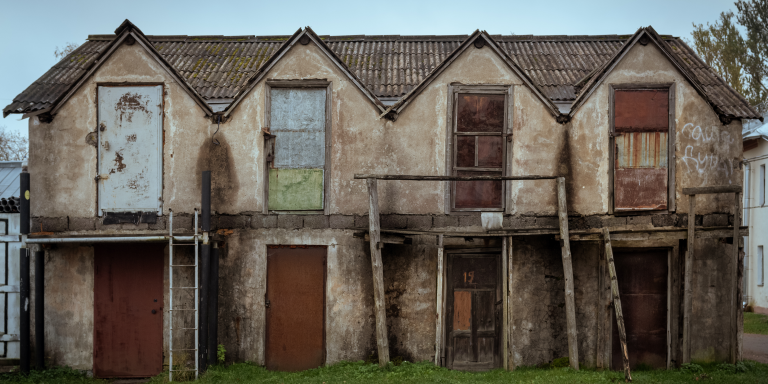 empty properties lined up. the facade is grey and decaying, and doors are boarded up and the buildings are in a state of disrepair.