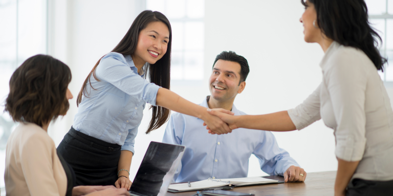 a group of professionals sitting around a table at a meeting. one person has a laptop, whilst two women shake hands across the table to signify the closing of a deal